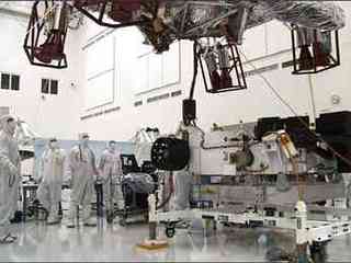 In this image, the car-size rover is in the middle of the picture with several team members surrounding it.  The team members are all dressed in special head-to-toe white suits, called 'bunny suits.'  One team member is holding on to a tether to guide the large insect-like descent stage down on top of the rover.  The descent stage looms high in this image.