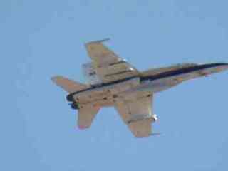 This image shows the underside of a grey F/A-18 jet with dark stripes flying against a blue sky from quite a distance away. Underneath the jet is a pod carrying sensors that measure the jet's temperature, pressure and vibration.