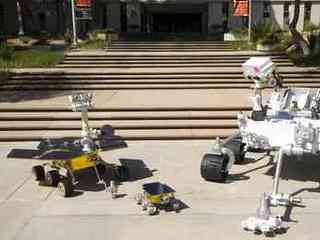 This image shows the front sides of three rovers viewed at an angle. On the right is the Mars Science Laboratory rover, which is the size of a small sport utility vehicle. Its 7-foot-long arm is extended in front of it, resting on the pavement. The camera 'eyes' at the top of its mast are turned downward at its smaller predecessors. In the middle, the tiny, wagon-size Sojourner rover has a deployable, alpha-particle X-ray spectrometer and single-color camera attached at its rear. On the left, the dune-buggy-size Mars Exploration Rover has its robotic arm extended in front of it, resting on the pavement. The panoramic camera 'eyes' at the top of its mast are turned upward, as if 'looking' at the bigger Mars Science Laboratory rover. All three rovers have six wheels, rocker-bogey suspension systems, rectangular bodies, solar panels, and antennas pointing upward from their spacecraft decks.