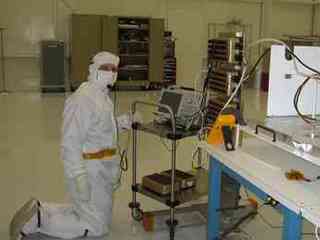This image was taken inside a large 'clean room,' where ATLO (Assembly, Testing, and Launch Operations) is taking place for the Mars Science Laboratory mission. In the foreground on the right are electronic components and to the left is a man leaning over the components. He is dressed in all white protective clothing, including a face mask and gloves.