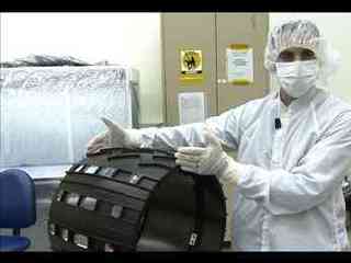 This image shows  engineer Sean Haggert. Wearing a white lab coat, gloves, face mask, and hair covering to protect spacecraft parts from contamination, engineer Sean Haggert places his hands on either edge of one of the black, cleated wheels of the Mars Science Laboratory rover.