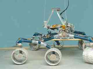 In this image, a large, six-wheeled rover sits on a concrete floor in a laboratory against a light-blue background.  The rover's 'legs,' or mobility system are a medium blue color and the large, cleated wheels are shiny silver.  On top of the mobility system is a silver-colored triangle that represents where the heart and brains of the rover will go when it is complete.