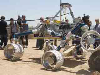 In this image, about a dozen people watch the large Scarecrow rover drive over a grayish orange boulder in an outdoor sandbox.  Scarecrow is about the size of a small compact car.  Its 'legs,' (mobility system) are a vibrant blue color and its large wheels are silver and covered in chevron-like cleats.  Members of the media hold cameras.