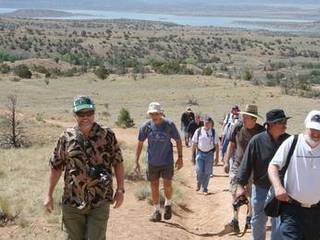 In this image, the Mars Science Laboratory Project Science Group, consisting of 11 men and 1 woman, are climbing up a dusty trail in New Mexico.  Wearing casual hiking clothes and hats, they are climbing towards the photographer of this image.  Behind them in the distance is a lake and mountains.