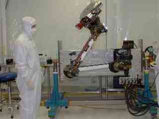 In this photo, two engineers from NASA Jet Propulsion Laboratory and Alliance Spacesystems are standing in a white room on either side of the Mars Science Laboratory rover's robotic arm.  They are dressed in white 'bunny suits,' and are covered from head to toe wearing white face masks to protect the equipment.  The arm is bent at the 'elbow' and raised high in the air about two feet higher than the engineer on the left.  The engineer on the right holds a control box that maneuvers the arm for testing.