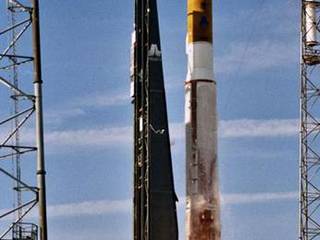 In this image, against a bright blue sky streaked with thin clouds, a Lockheed Martin-built gold and white rocket lifts off its pad at Cape Canaveral Air Station, Florida. Fire bursts from the bottom of the rocket which is surrounded by four lightning suppression towers, two of which are in view in this picture.