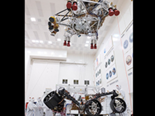 Engineers and technicians working on the Mars 2020 spacecraft look on as a crane lifts the rocket-powered descent stage away from the rover after a test.