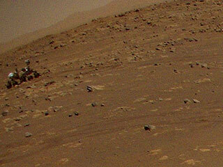 NASA’s Perseverance Mars rover is visible in the upper left corner of this image the agency’s Ingenuity Mars Helicopter took during its third flight, on April 25, 2021. The helicopter was flying at an altitude of 16 feet (5 meters) and roughly 279 feet (85 meters) from the rover at the time.