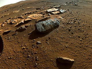 This is a colored image of Perseverance capturing the sandy, rocky surface of Mars. Perseverance’s wheel in the bottom left of the image. There are large boulders and tiny rock particles present in this image.  