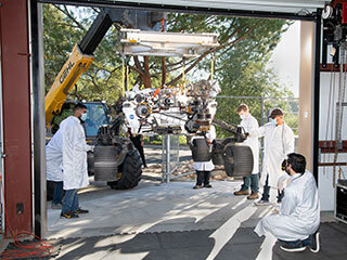 On Oct. 29, 2021, a heavy-duty vehicle transports the Perseverance rover’s engineering model, called OPTIMISM, from a test lab to the Mars Yard garage at JPL.