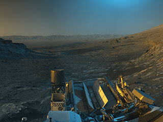 NASA’s Curiosity Mars rover used its black-and-white navigation cameras to capture panoramas of this scene at two times of day. Blue, orange, and green color was added to a combination of both panoramas for an artistic interpretation of the scene.