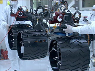 In the middle of this image, three wheels are shown raised by a lift, with engineers on both sides of the wheels in the cleanroom, where the Curiosity rover is being assembled. The wheels are made out of aluminum but have a black coating, which prevents the wheels from slipping.  The wheels are attached to dark poles, which bend like 'joints.'  Several black and red cable wires are visible coming out of the poles.  An engineer stands in the background holding a clipboard and looking closely at the rover.  A desk and various tools are visible behind the wheels on the right.