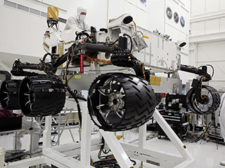 This image was taken in the clean room where the Curiosity rover is being assembled.  It shows the rover, which is about the size of an SUV, hoisted on a white lift, with its black wheels suspended in the air.   One engineer is on top of the hoist and is leaning over the rover body, while another is looking up on the ground floor to the right of the rover.  Both engineers are wearing white "bunny suits" to keep them from contaminating any equipment.