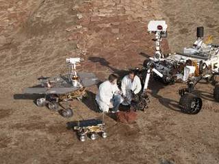 Two spacecraft engineers join a grouping of vehicles providing a comparison of three generations of Mars rovers developed at NASA's Jet Propulsion Laboratory, Pasadena, Calif. The setting is JPL's Mars Yard testing area.