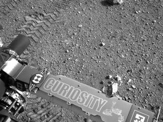 This image shows a close-up of track marks from the first test drive of NASA's Curiosity rover. The rover's arm is visible in the foreground.