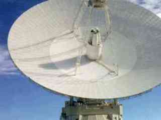 The image shows a very tall, wide, dish-shaped device that is about three times as wide and as tall as the two-story dome that supports it. Four metal ladderlike extensions form a pyramidal structure topped by a box-like frame used for transmission and reception. The entire array is pointed skyward. In the background is a partly cloudy, blue sky above sagebrush-covered desert hills.