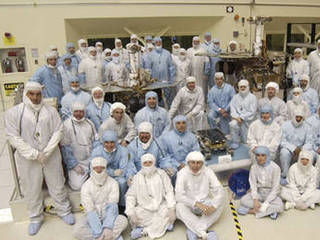 A team of about 50 men and women in white and blue cleanroom smocks and bonnets  stand and sit around their creations: Spirit and Opportunity.  The twin rovers of the Mars Exploration Rover mission are about the size of golf carts.  The significantly smaller, microwave-sized Sojourner rover from the 1997 Pathfinder mission sits in the middle.