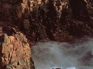 This image shows a white, frothy stream flowing down a canyon between tall, red, volcanic cliffs.