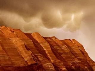 This image shows layered, red cliffs rising up from a flat, brown surface. The cliff surface slants from right to left, with the nearest cliffs on the right-hand side of the image and those farther away toward the left. Pinkish-tan clouds hover above the crests of the cliffs.